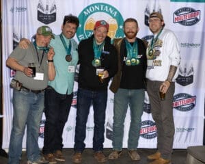 Five men wearing medals pose together on a stage in front of a sponsor banner, each holding drinks and smiling.