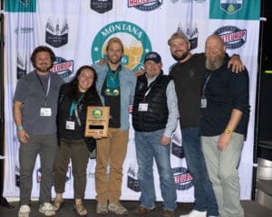 Six adults posing together at a trophy ceremony in front of a banner with Montana beer awards logos and sponsor labels.