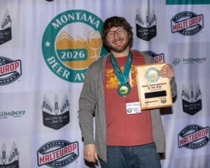 Smiling man with glasses and a beard, wearing a medal, holding a wooden Montana Beer Awards plaque at a beer?event backdrop.