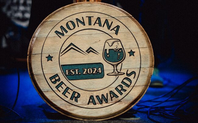 Wooden circular sign reading Montana Beer Awards with a beer glass illustration, set against a dark stage backdrop (EST. 2024).